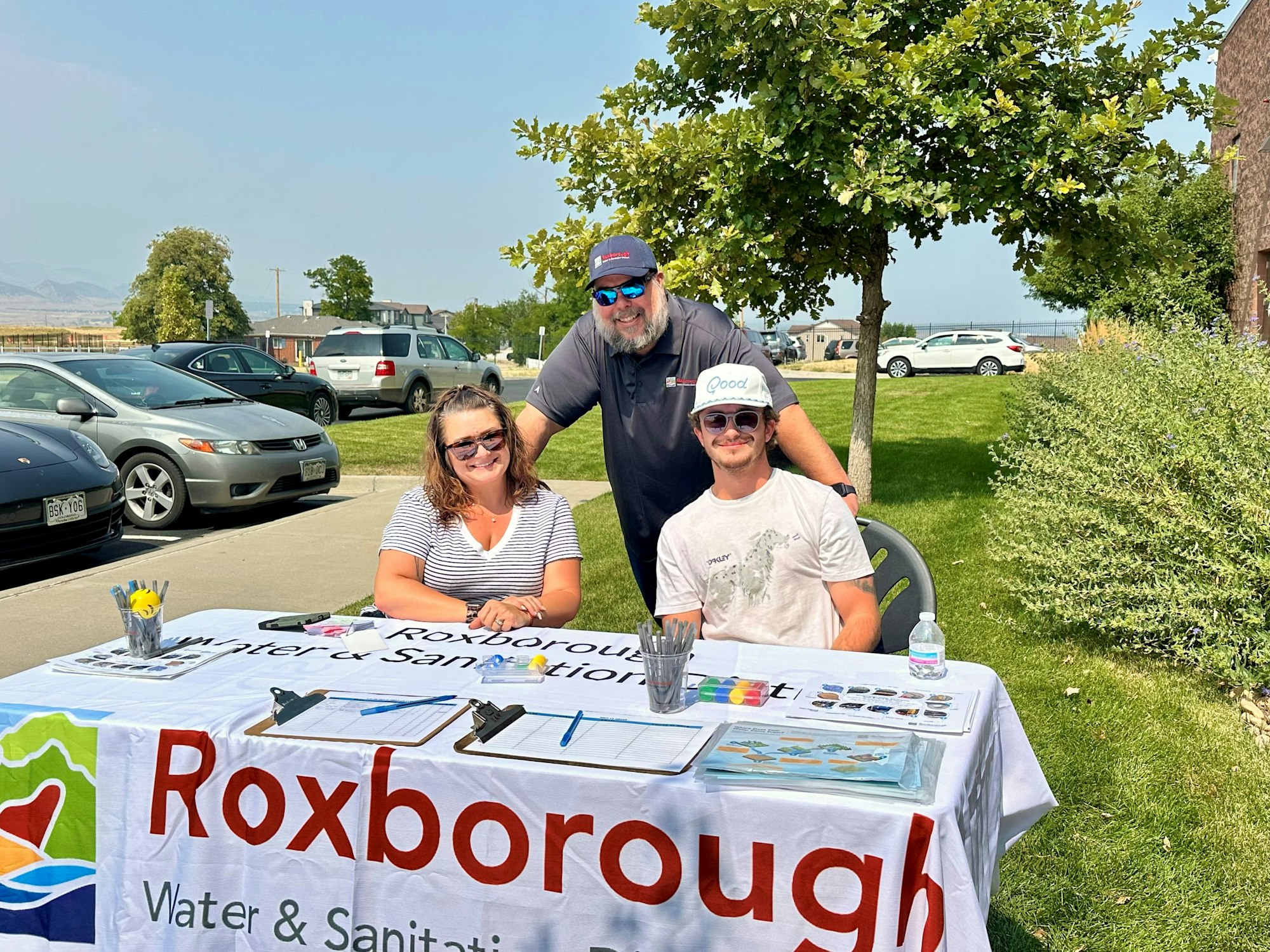 Three people at an outdoor booth for Roxborough Water & Sanitation, with clipboards and pens on a table.