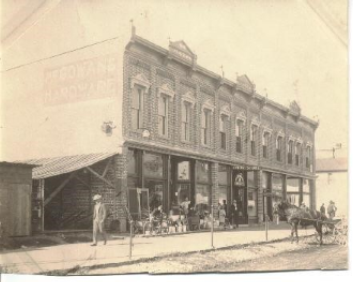 A vintage photo of a hardware store with people, a horse-drawn carriage, and classic architecture.
