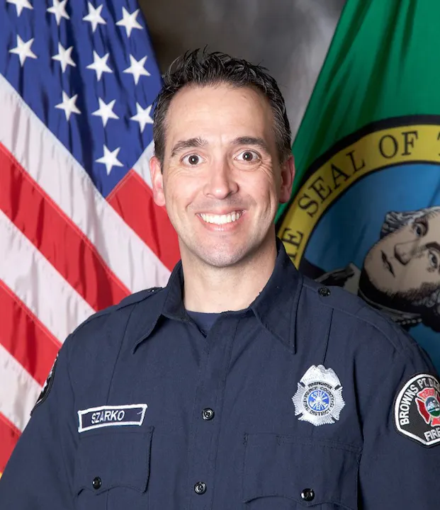 The image shows a smiling individual in a dark uniform with insignia, standing in front of U.S. and state flags.