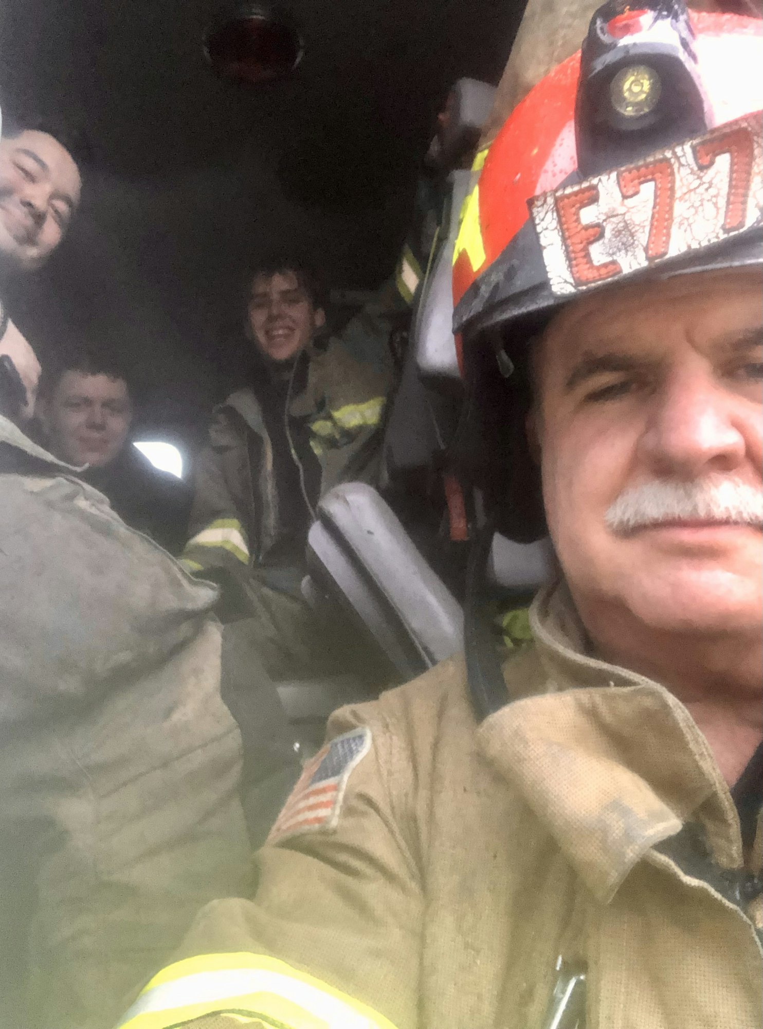Face shot of fire captain in bunker gear in the cab of a fire engine, with firefighting crew behind him.