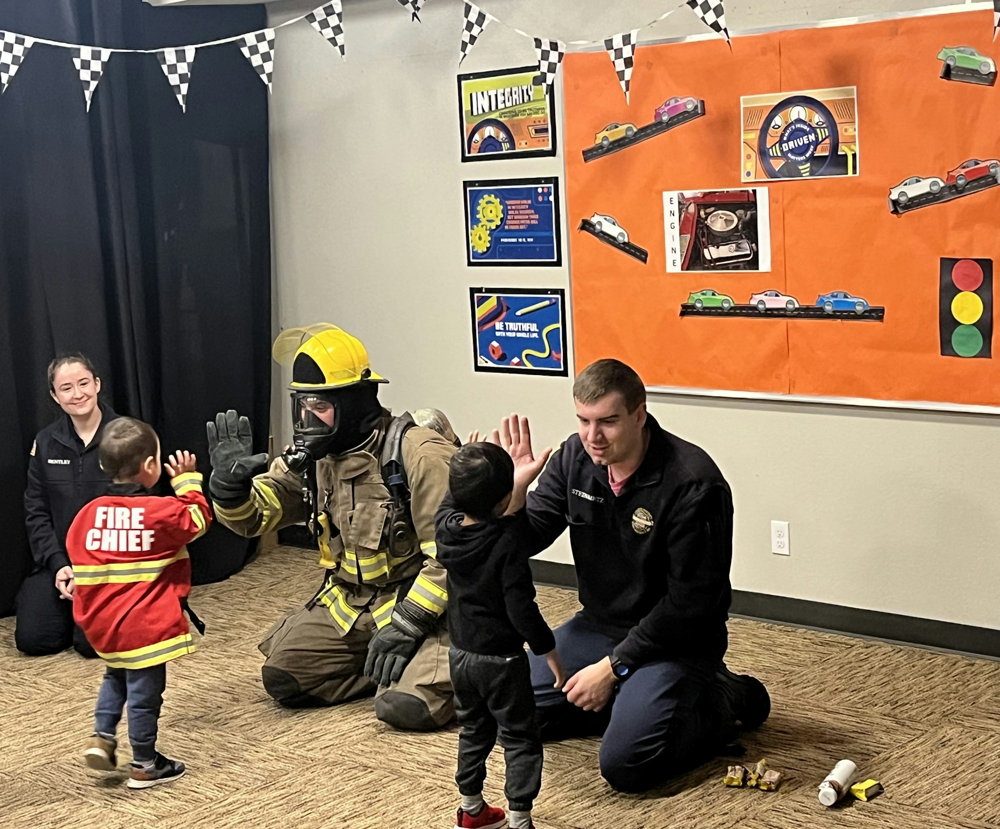 Two male and one female firefighter, kneeling, providing fire safety instruction to two preschool boys