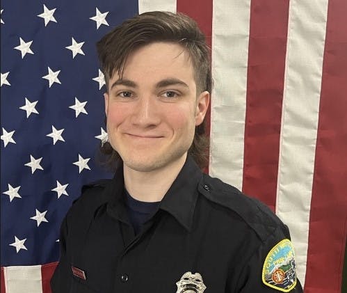 A smiling person in a uniform with a badge in front of an American flag.
