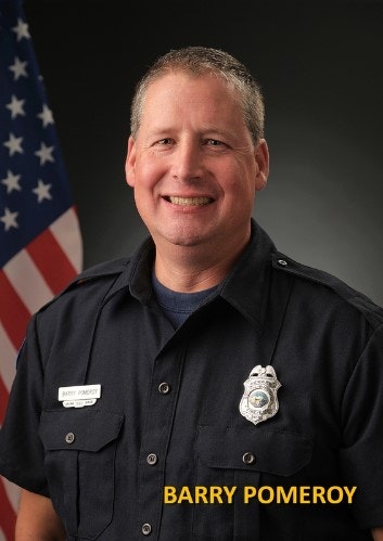 A smiling person in a uniform with a badge in front of an American flag.
