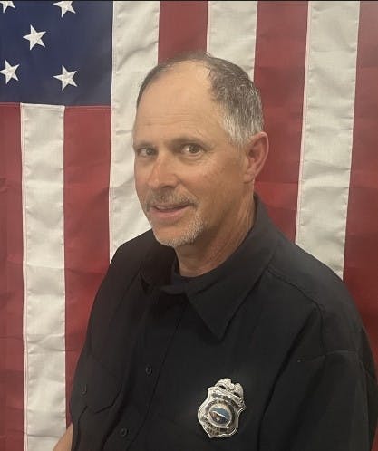A man in a black shirt with a badge, in front of an American flag.