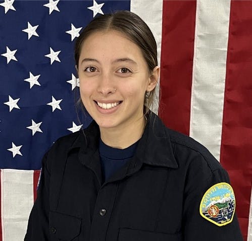 A smiling person in uniform with a badge in front of an American flag.