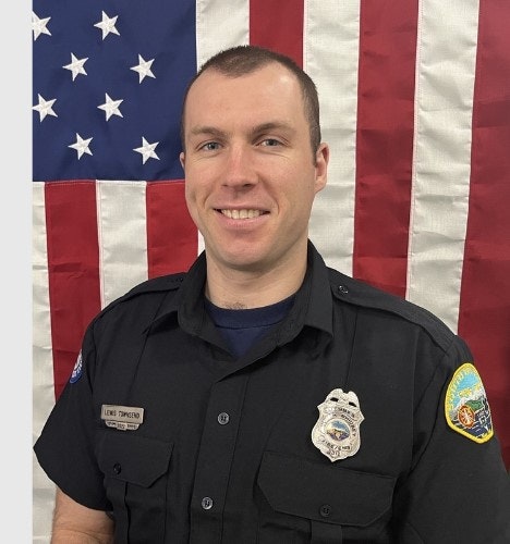 A smiling police officer in uniform in front of an American flag.