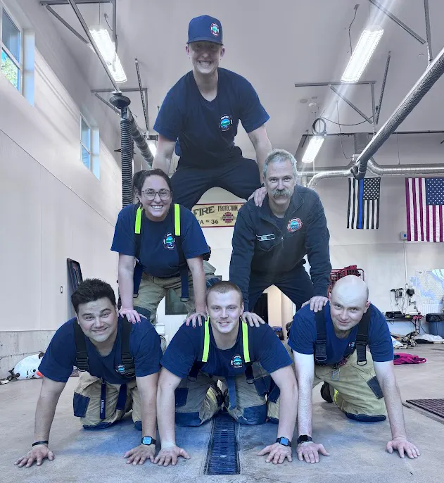 A group of six firefighters is posing in a human pyramid in a fire station, showcasing teamwork and camaraderie.