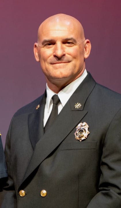 A person in a formal uniform with badges, smiling against a plain background.