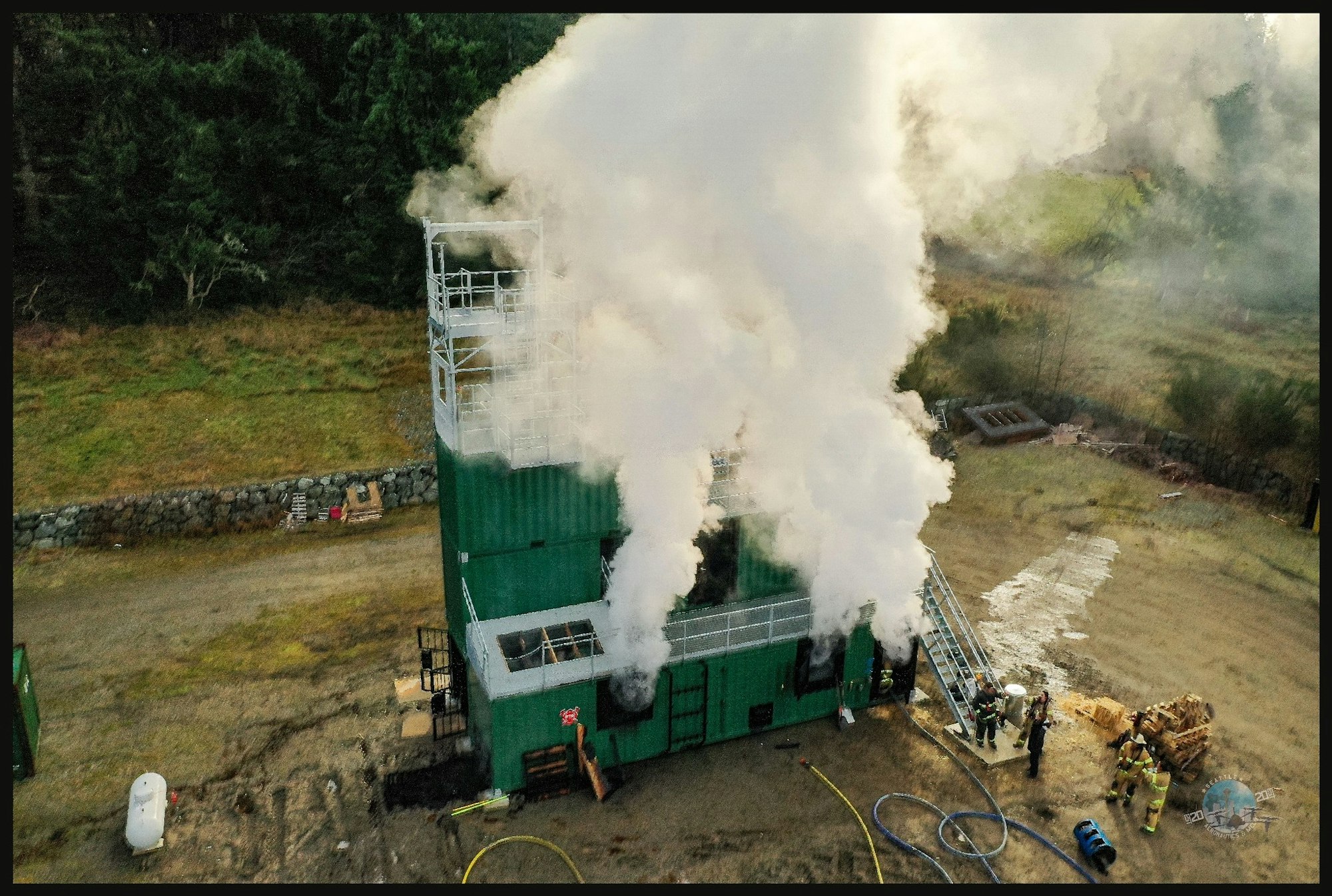 Smoke billows from a green training building, while firefighters operate nearby, likely conducting fire training exercises.