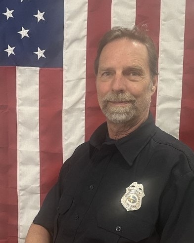 A man in a black uniform with a badge in front of an American flag.