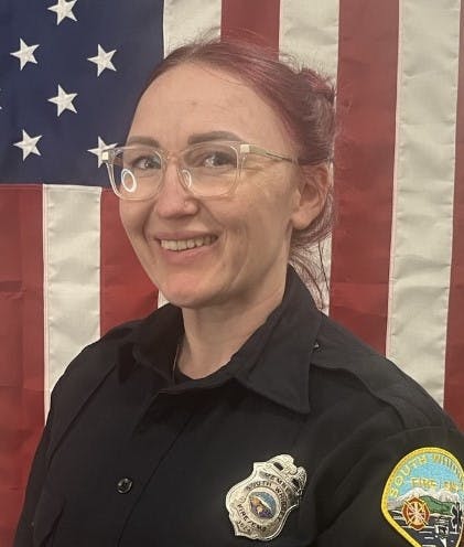 A smiling person in uniform with a badge, in front of the American flag.