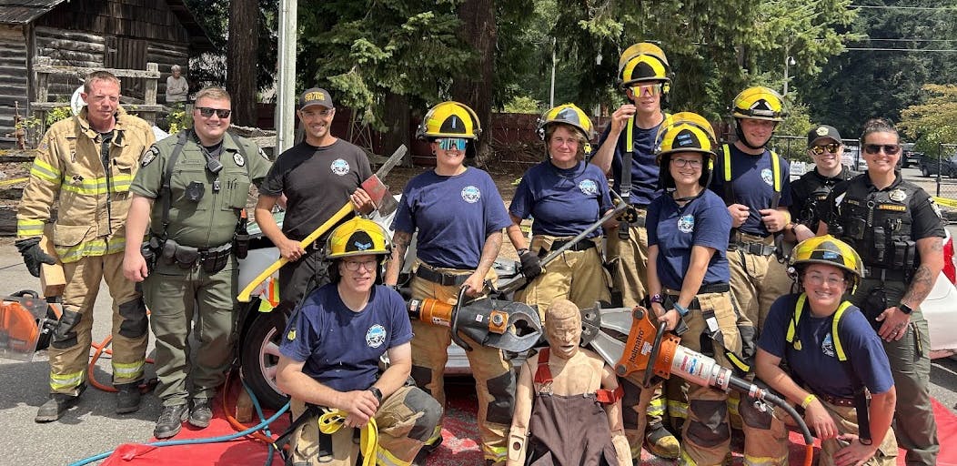 A group of firefighters and officers pose outdoors with rescue equipment and a training dummy.