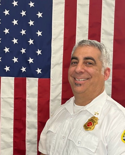 A smiling person in a white uniform with a badge in front of an American flag.
