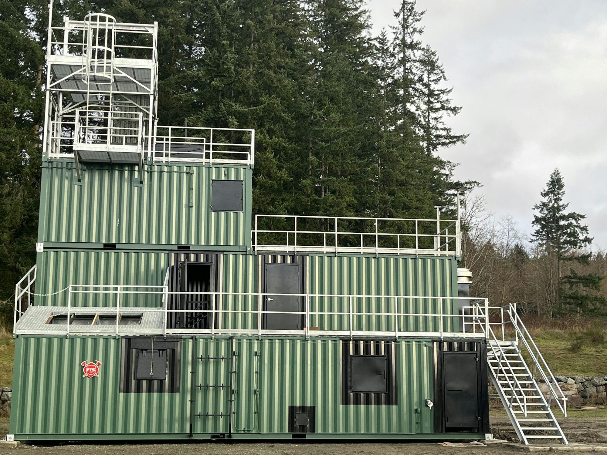 The image shows a green, multi-level training facility made from shipping containers, set against a forest backdrop.