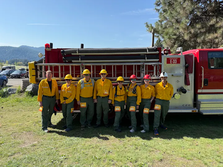 A group of people in yellow uniforms stand in front of a red fire truck outdoors.