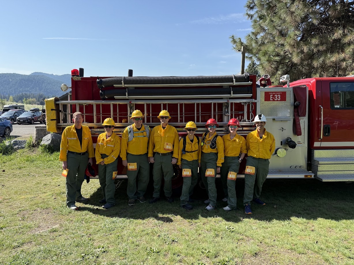 A group of people in yellow uniforms stand in front of a red fire truck outdoors.