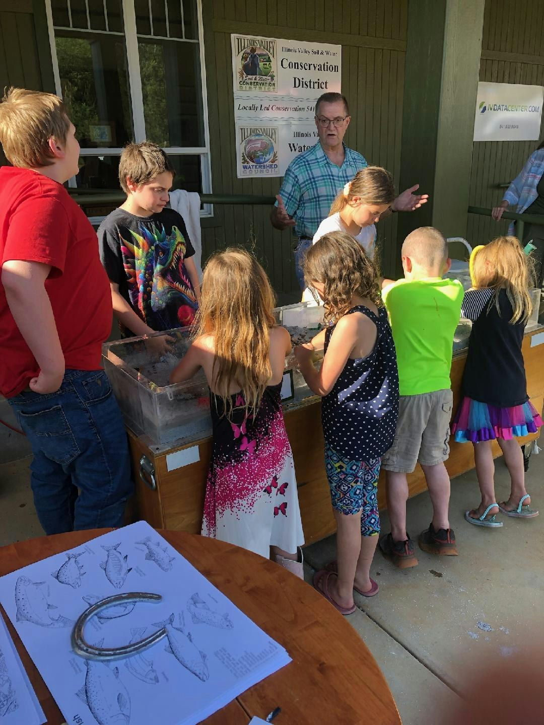 Children engaged in an educational activity outdoors, with a man presenting and conservation signage in the background.
