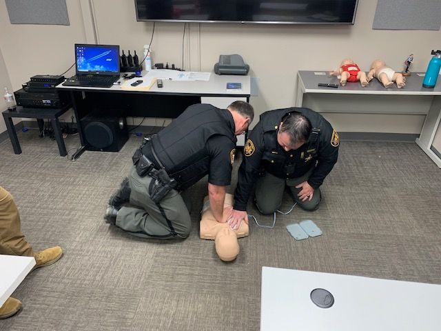 Two people practicing CPR on a mannequin in a training room with medical equipment and computers.