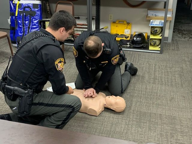 Two uniformed individuals practicing CPR on a training dummy in a room with equipment.