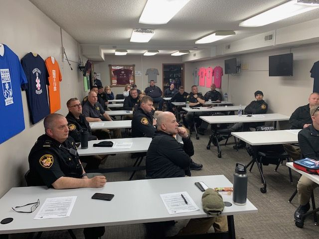 A group of uniformed officers sits in a classroom setting with shirts on the wall.
