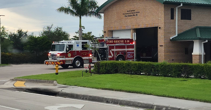 A fire truck is parked outside a fire station, with greenery and a pleasant sky in the background.