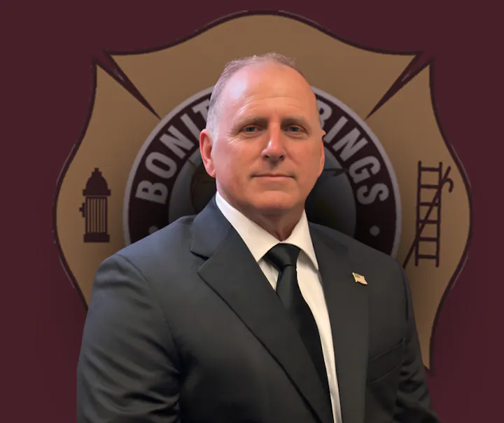 A man in a suit stands in front of a badge emblem related to a fire department or similar organization.