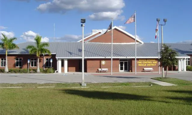 An exterior view of a brick building with the sign "Fire Control and Rescue District," featuring flags and palm trees.