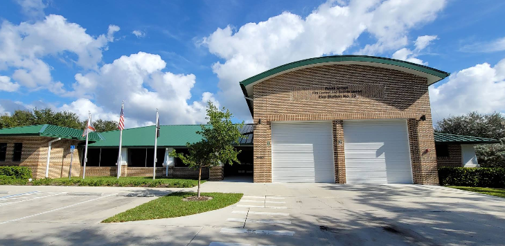 A brick building with a green roof and two garage doors, surrounded by trees and parked vehicles, under a blue sky.