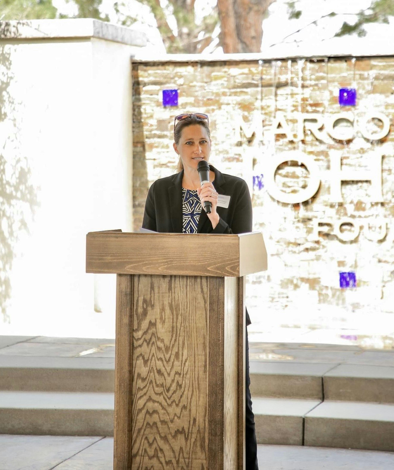 A person speaking at a wooden podium outdoors, with a decorative wall behind them.