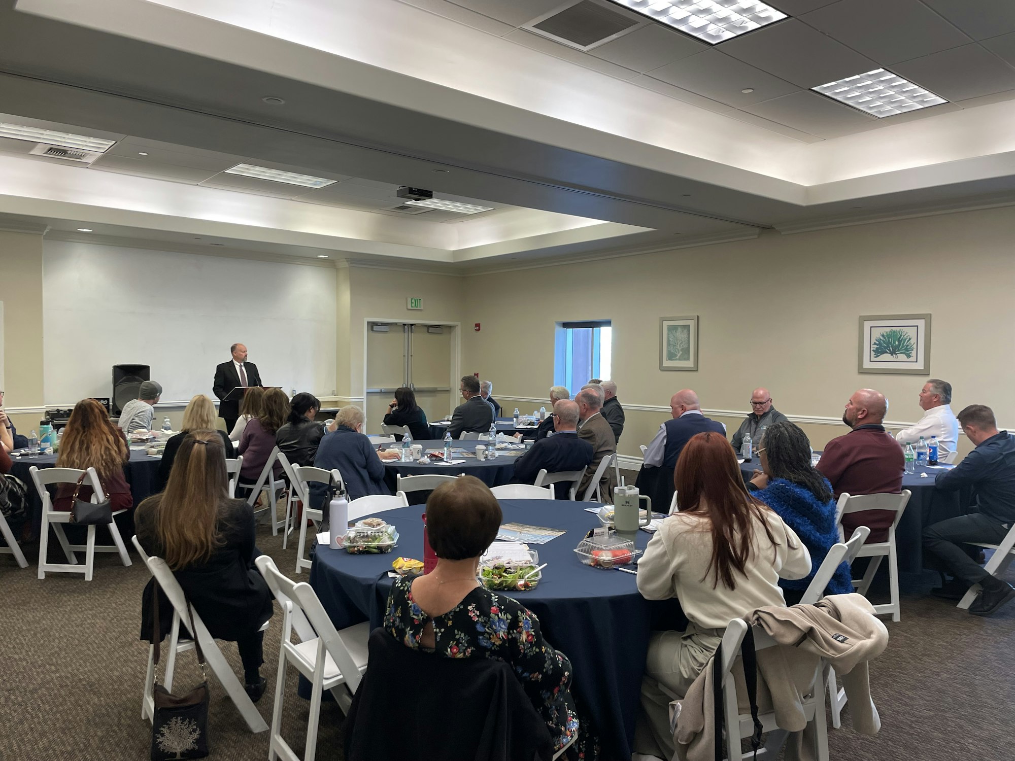 A group of people sitting at tables in a conference room, listening to a speaker at a presentation or meeting.