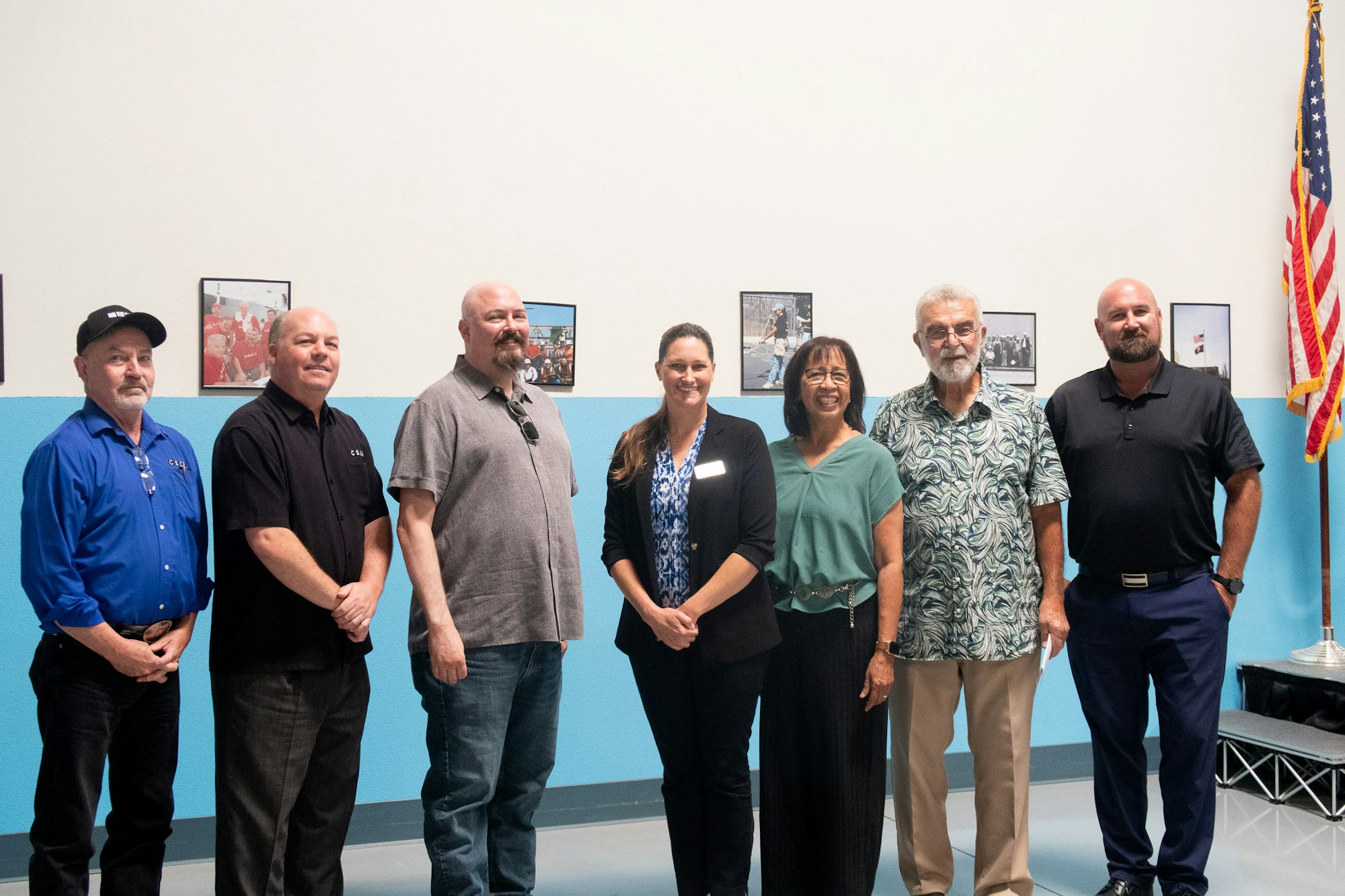 A group of seven people stands together in a room, with framed photos on the wall and an American flag nearby.