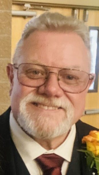 A man with glasses and a white beard, wearing a suit and tie, with a yellow flower boutonnière.