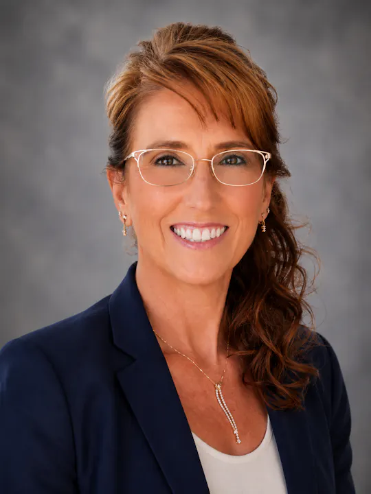 A professionally dressed woman with glasses smiles in front of a neutral background, showcasing her confidence and approachability.