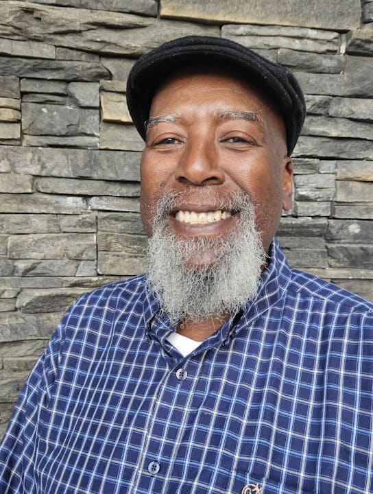 A person with a beard and a plaid shirt smiles in front of a stone wall.