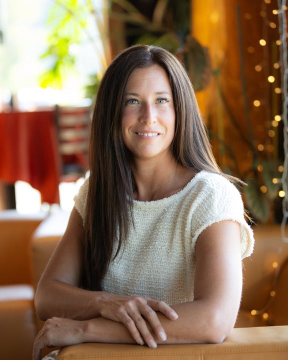 A woman with long hair, wearing a textured white top, is seated indoors, smiling with arms crossed. Blurred background has warm lighting.