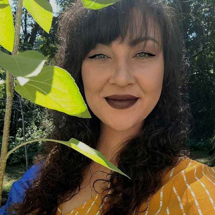 A person with curly hair and dark lipstick, smiling near green leaves outdoors.