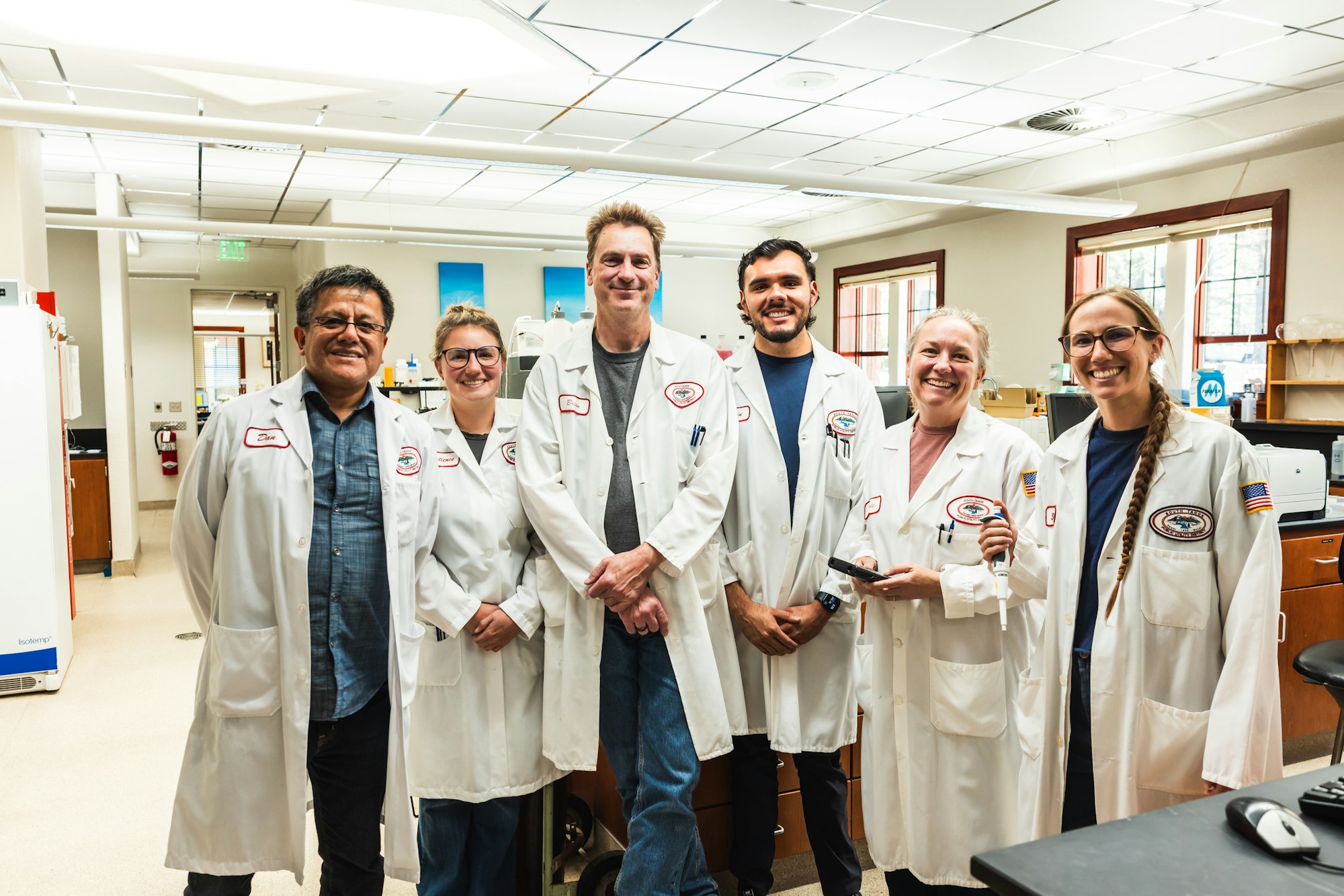 A group of six individuals in lab coats smiling together in a laboratory setting.