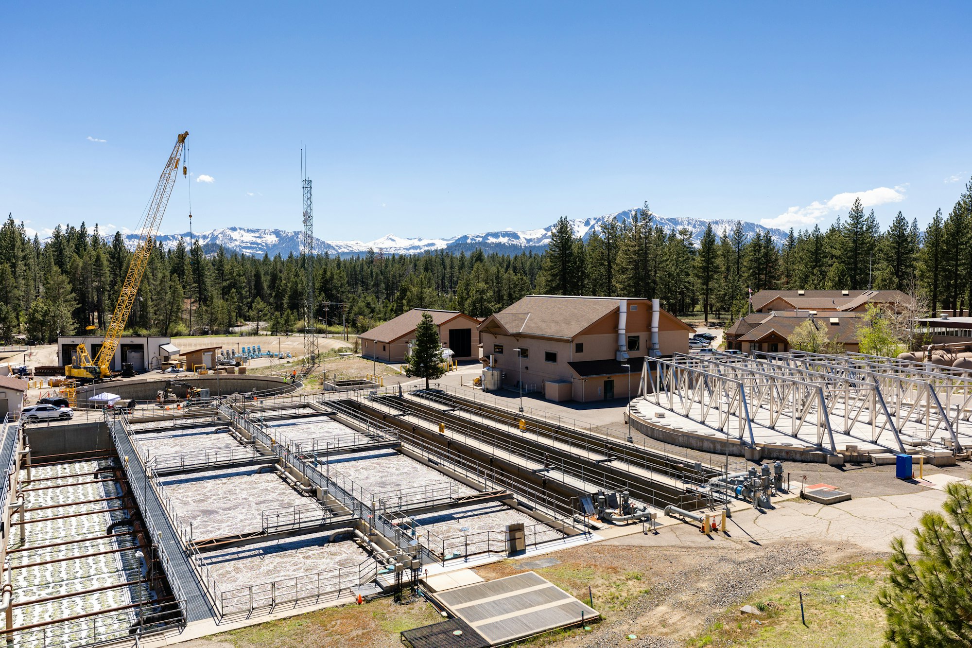 A water treatment plant with basins, buildings, and a crane, set against a backdrop of mountains and trees.