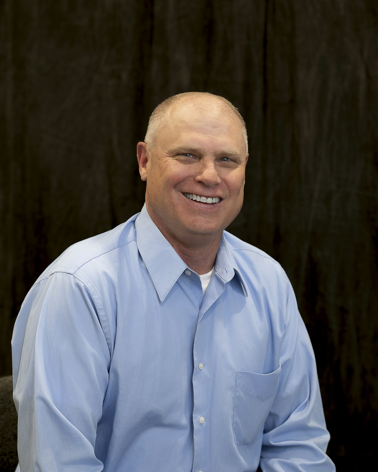 A smiling man in a light blue shirt against a dark background.