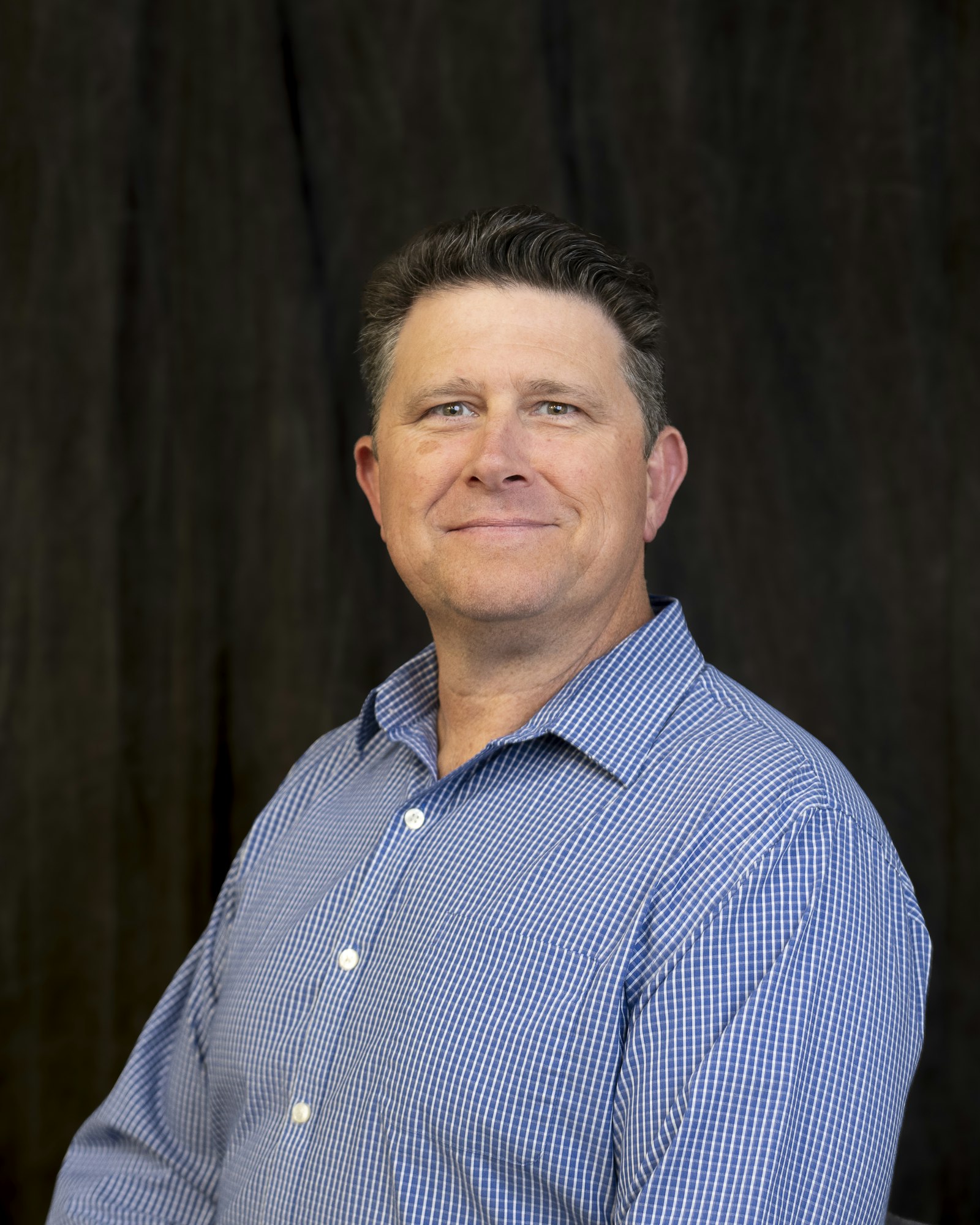 A person wearing a blue checkered shirt, standing against a dark background.