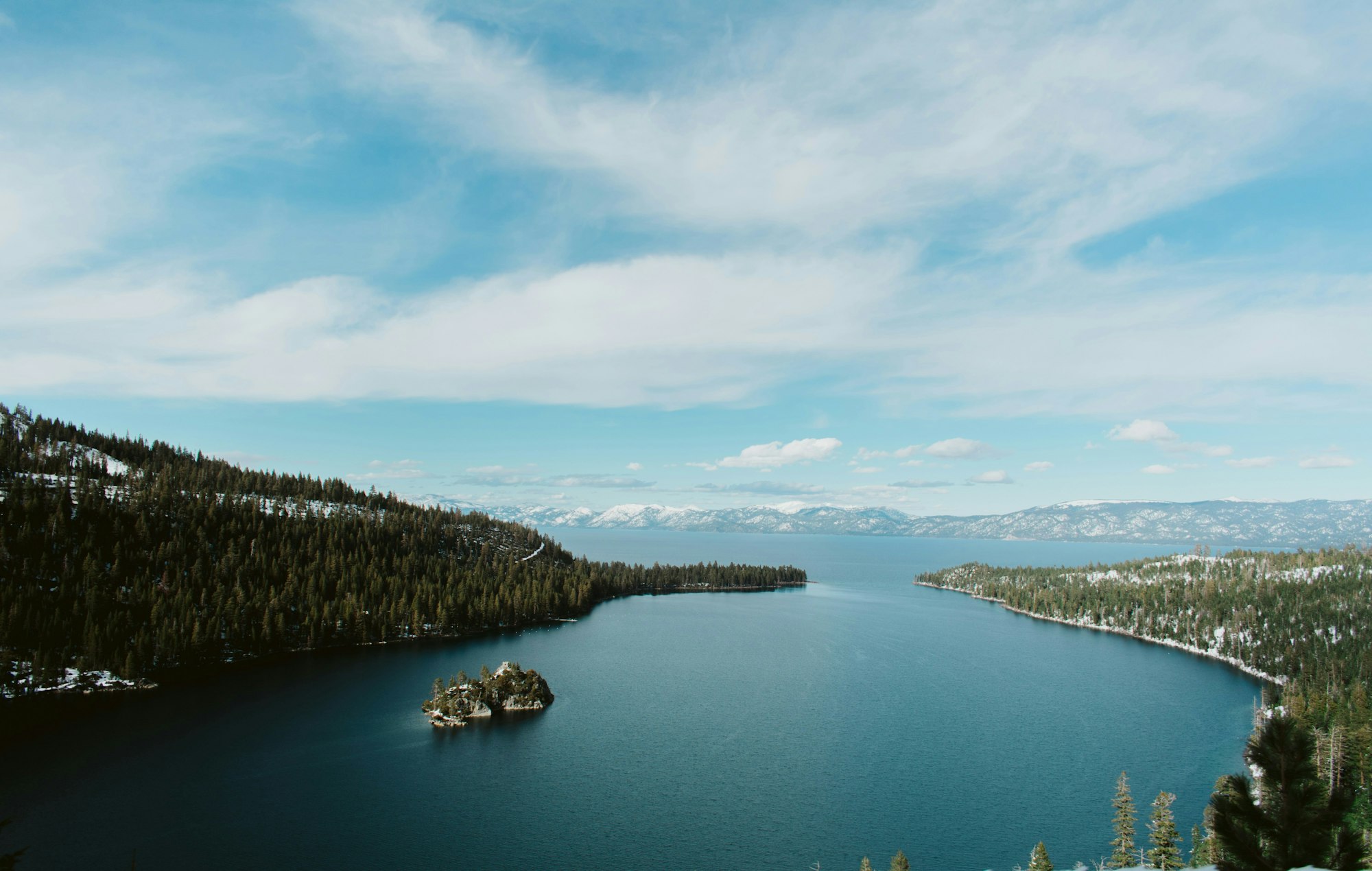 A serene lake bordered by forest with snowy mountains in the background under a partly cloudy sky.