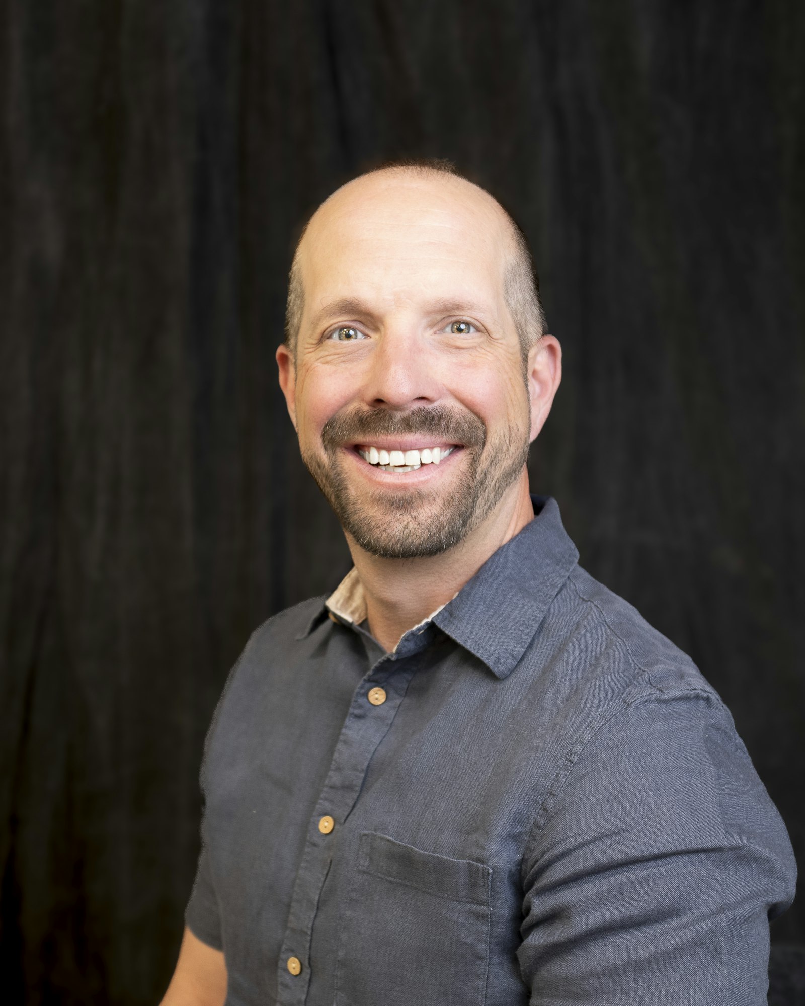 A smiling man in a blue shirt against a dark background.