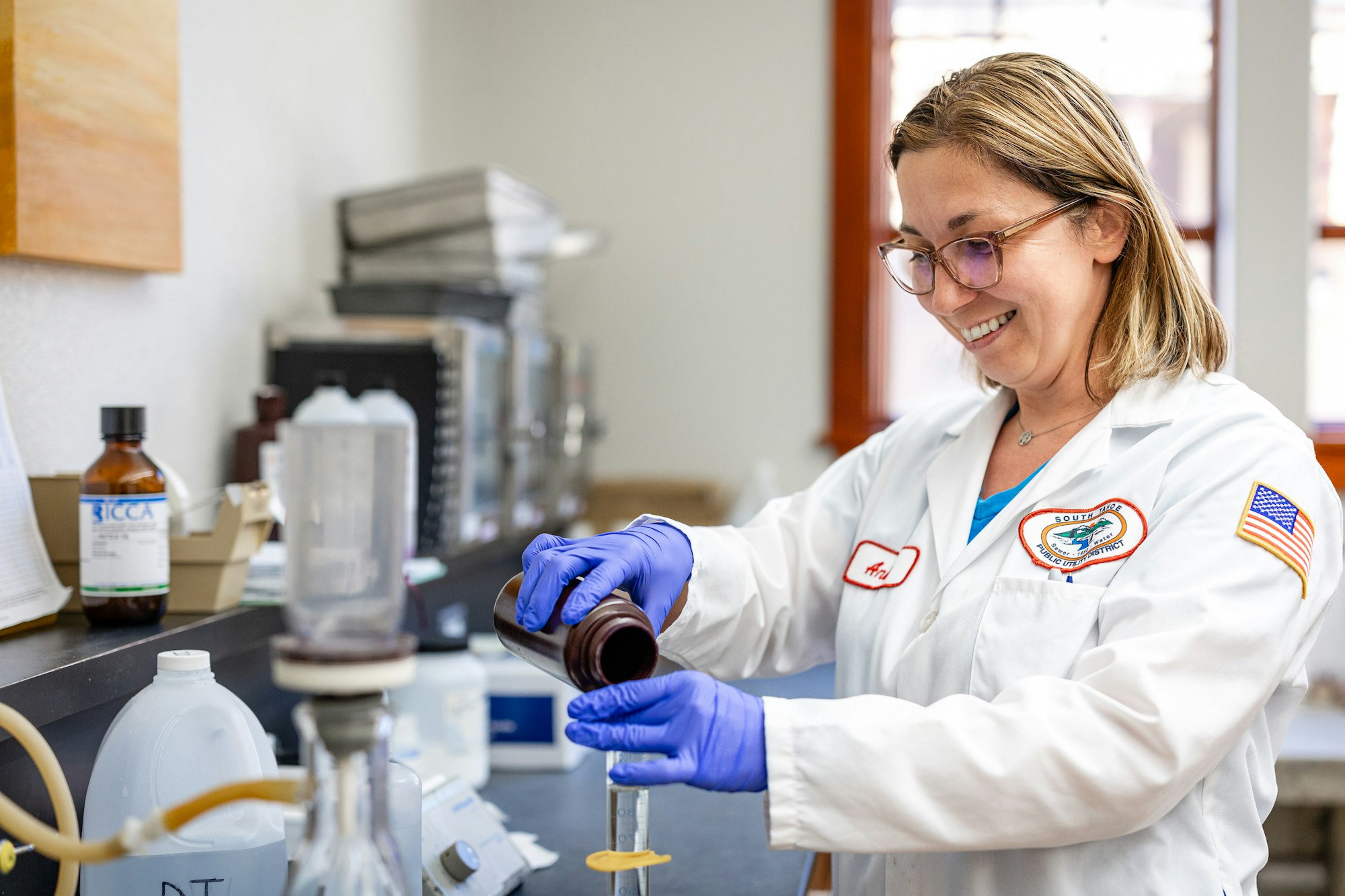 A smiling person wearing a lab coat and gloves is handling a bottle and a syringe in a lab setting.