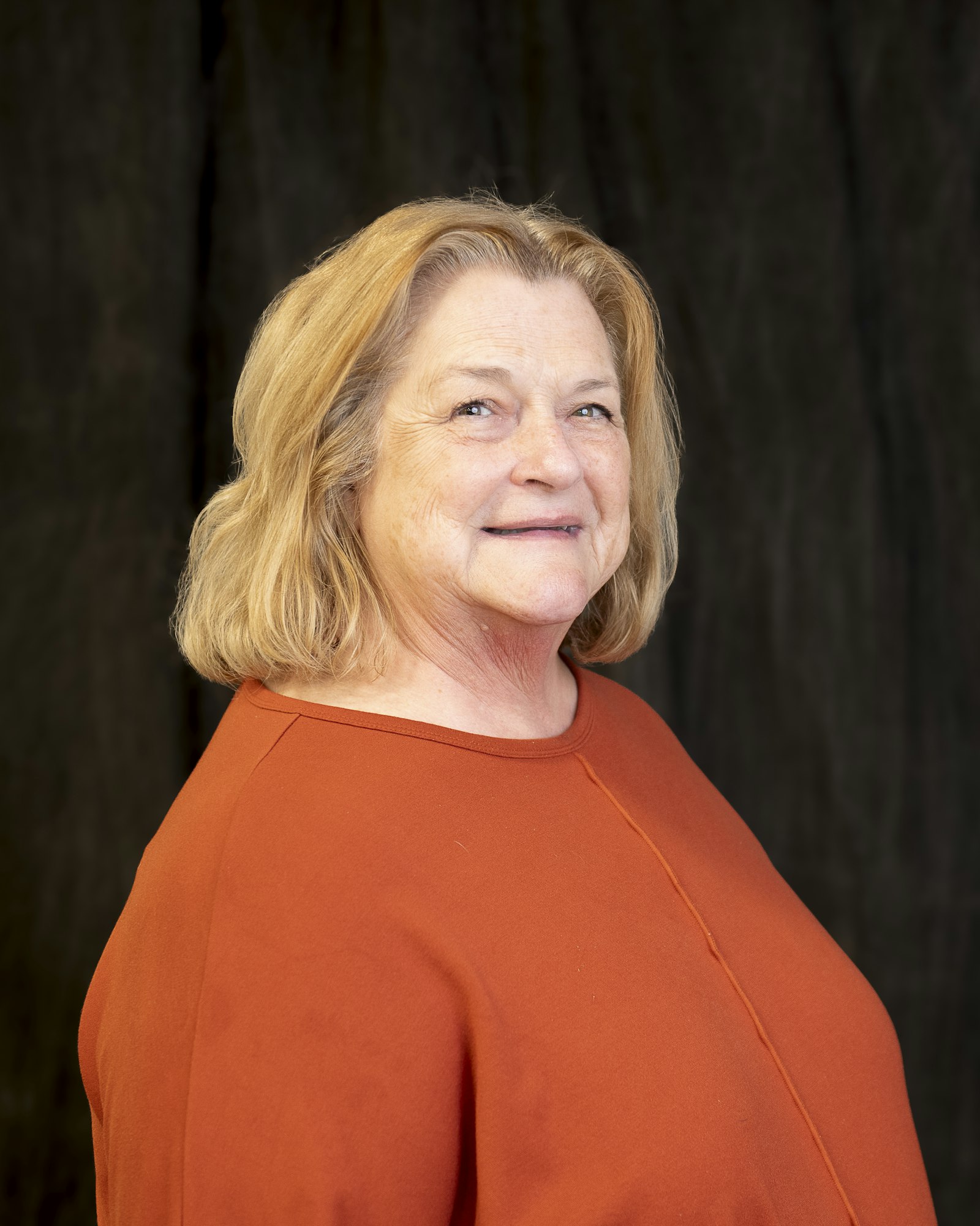 An older woman with blonde hair, wearing an orange top, smiling against a dark background.
