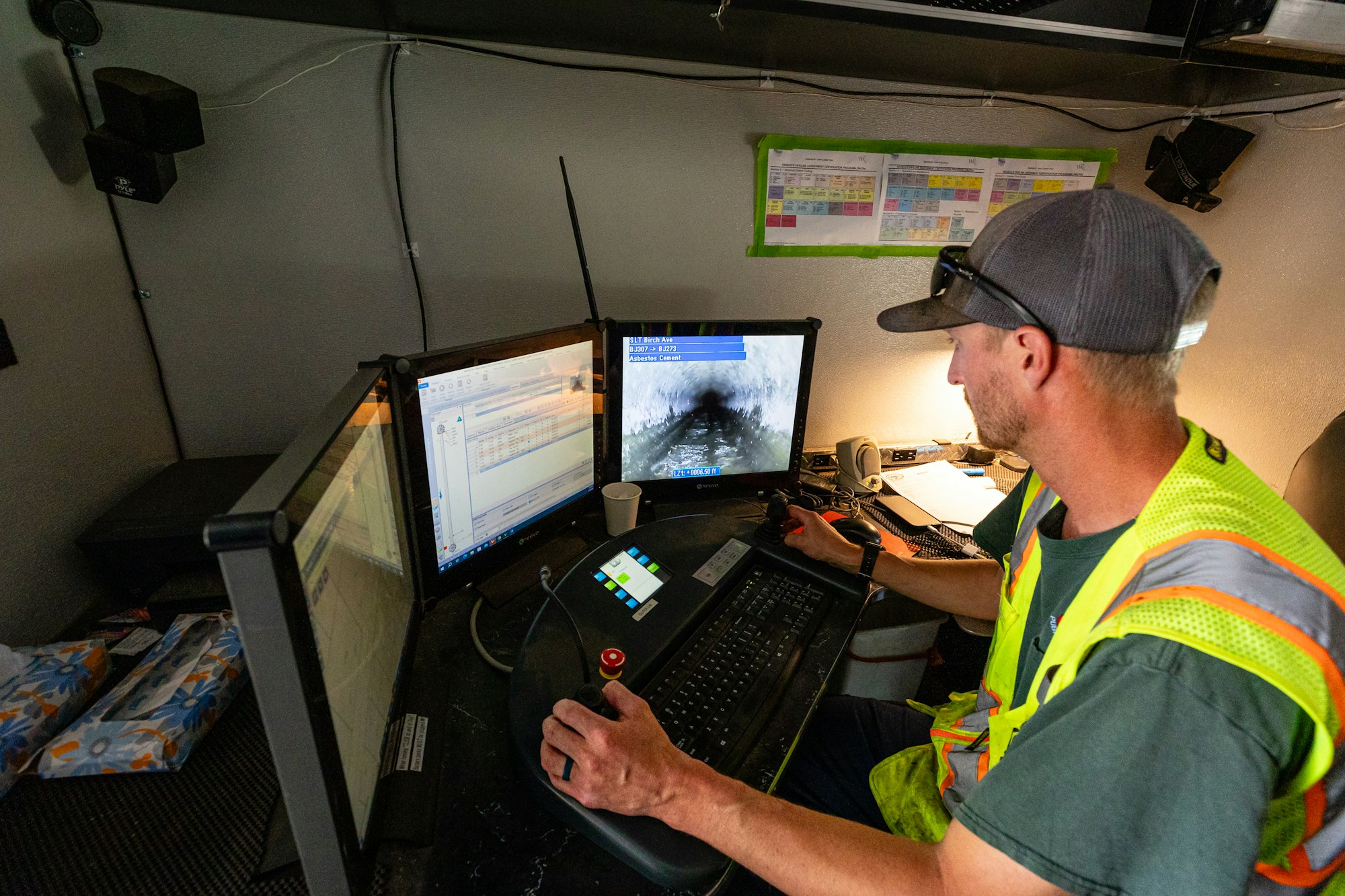 A sewer crew worker in a cap and safety vest at a desk with multiple monitors, one displaying a camera feed from sewer line, operating a joystick.
