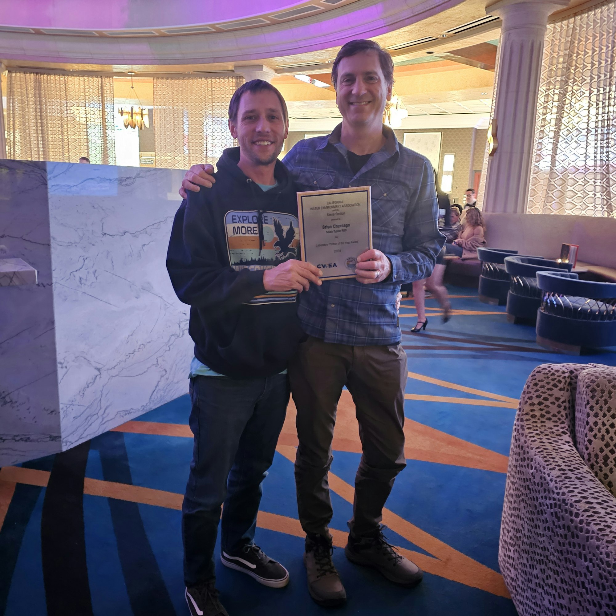 Two people are standing together indoors, smiling, with one holding a certificate.