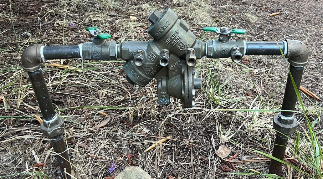 A metal water control valve assembly with multiple joints and two green handles, set in a grassy area.