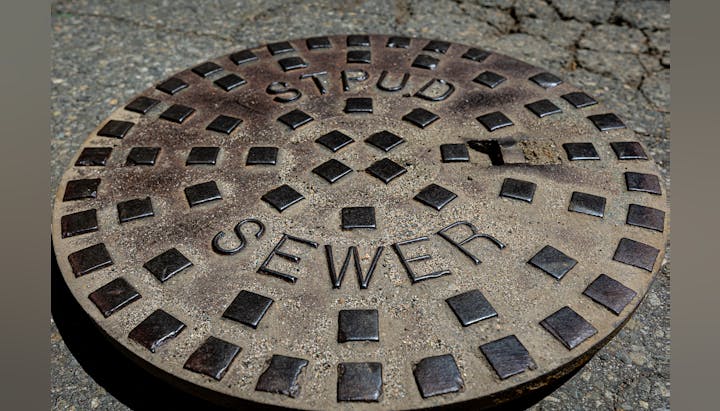 A close-up of a round, rusty manhole cover with the word "SEWER" on it.