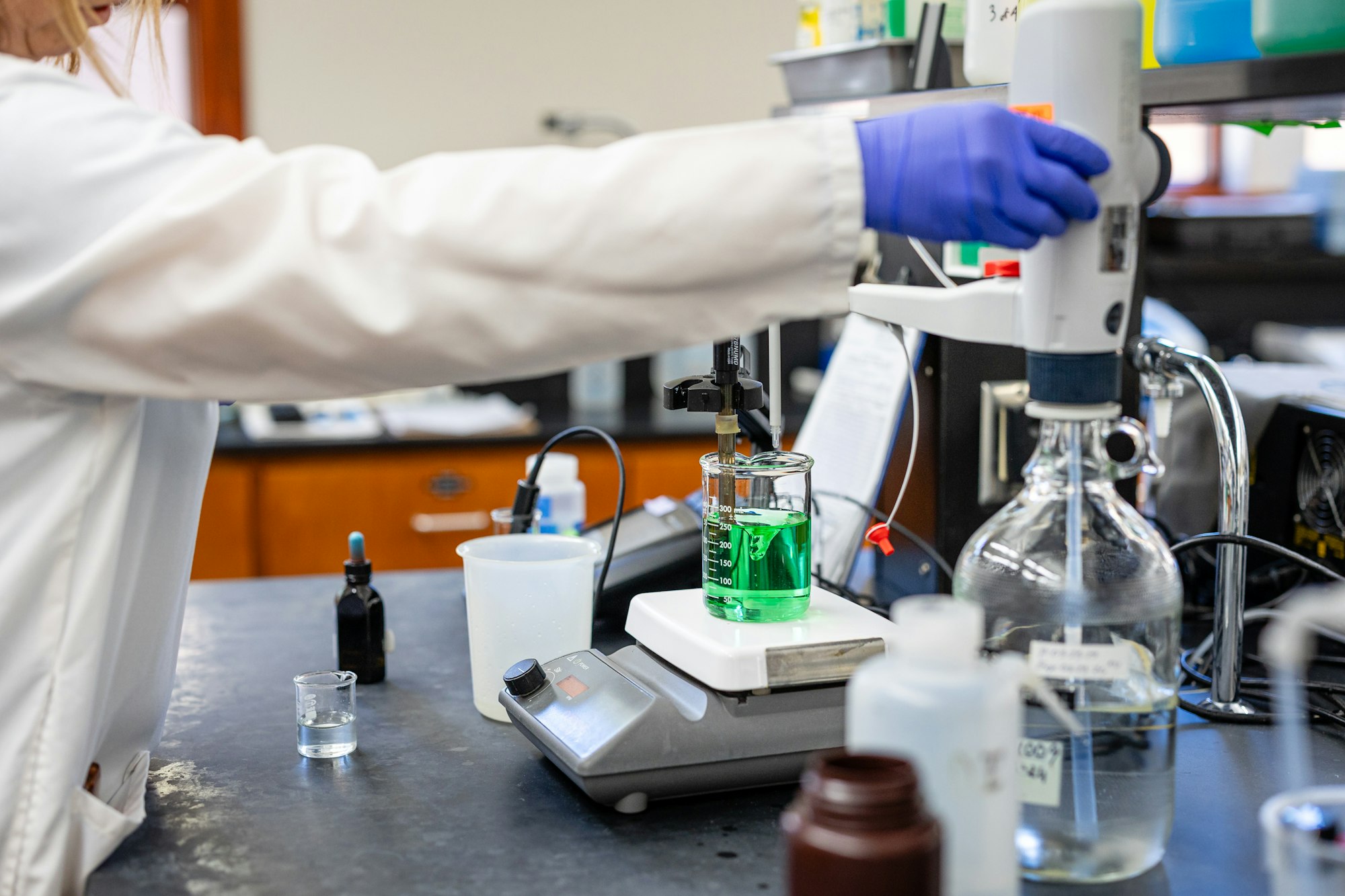 A person pipetting a green liquid in a chemistry lab setup.