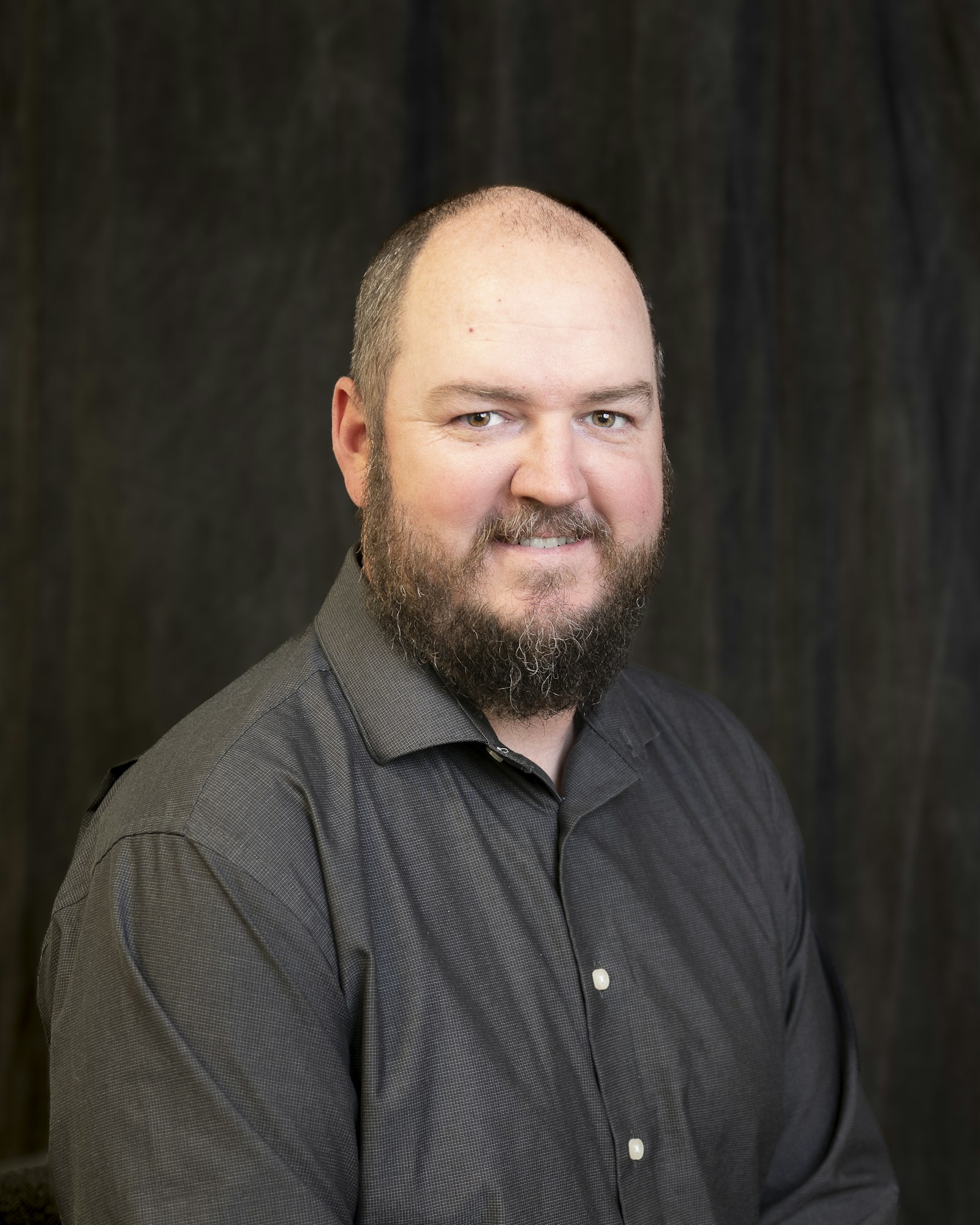 A man with a beard and a dark shirt, smiling in front of a dark background.